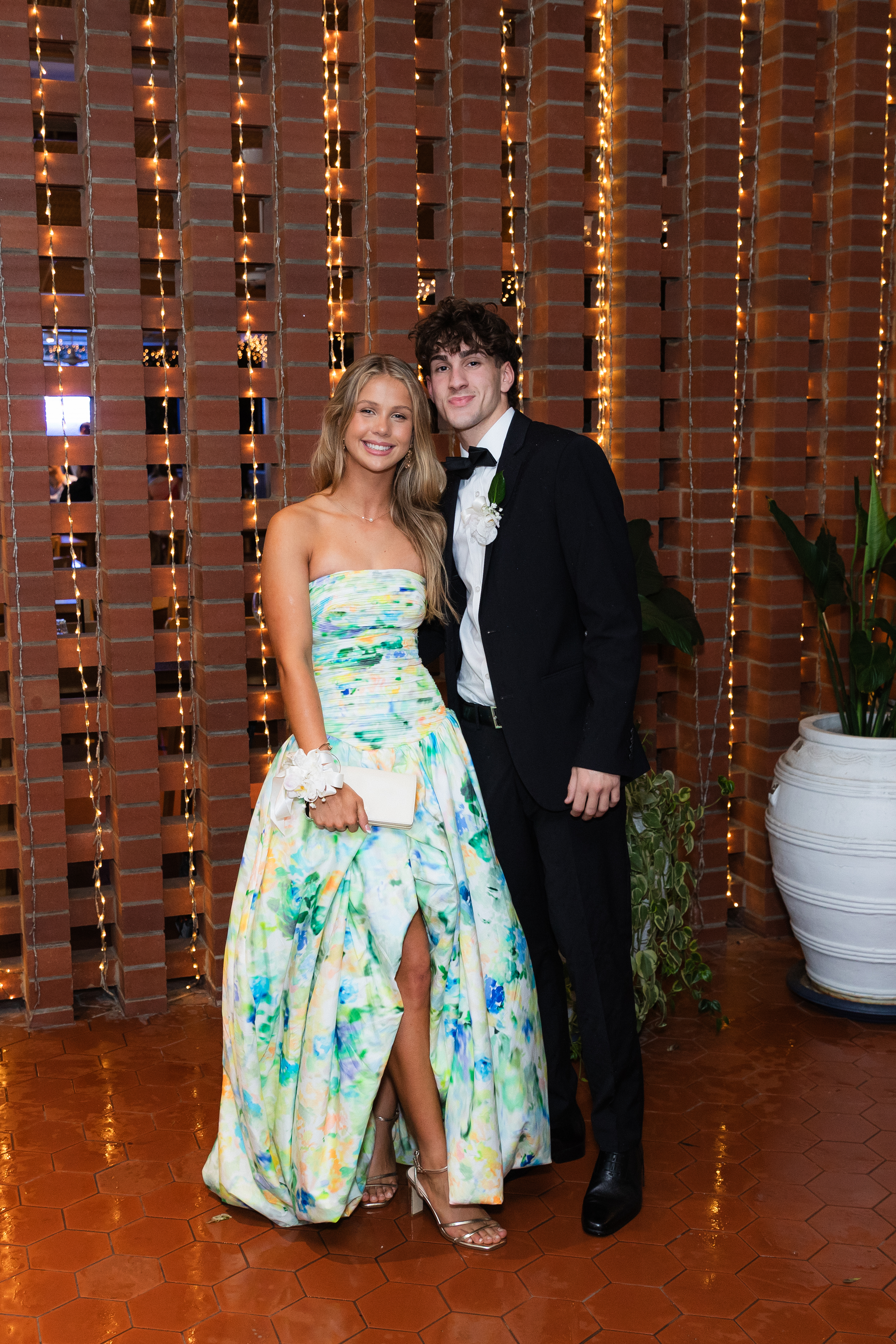 Two people posing together in formal attire at an indoor event, standing in front of a brick wall with hanging string lights, photographed full length.