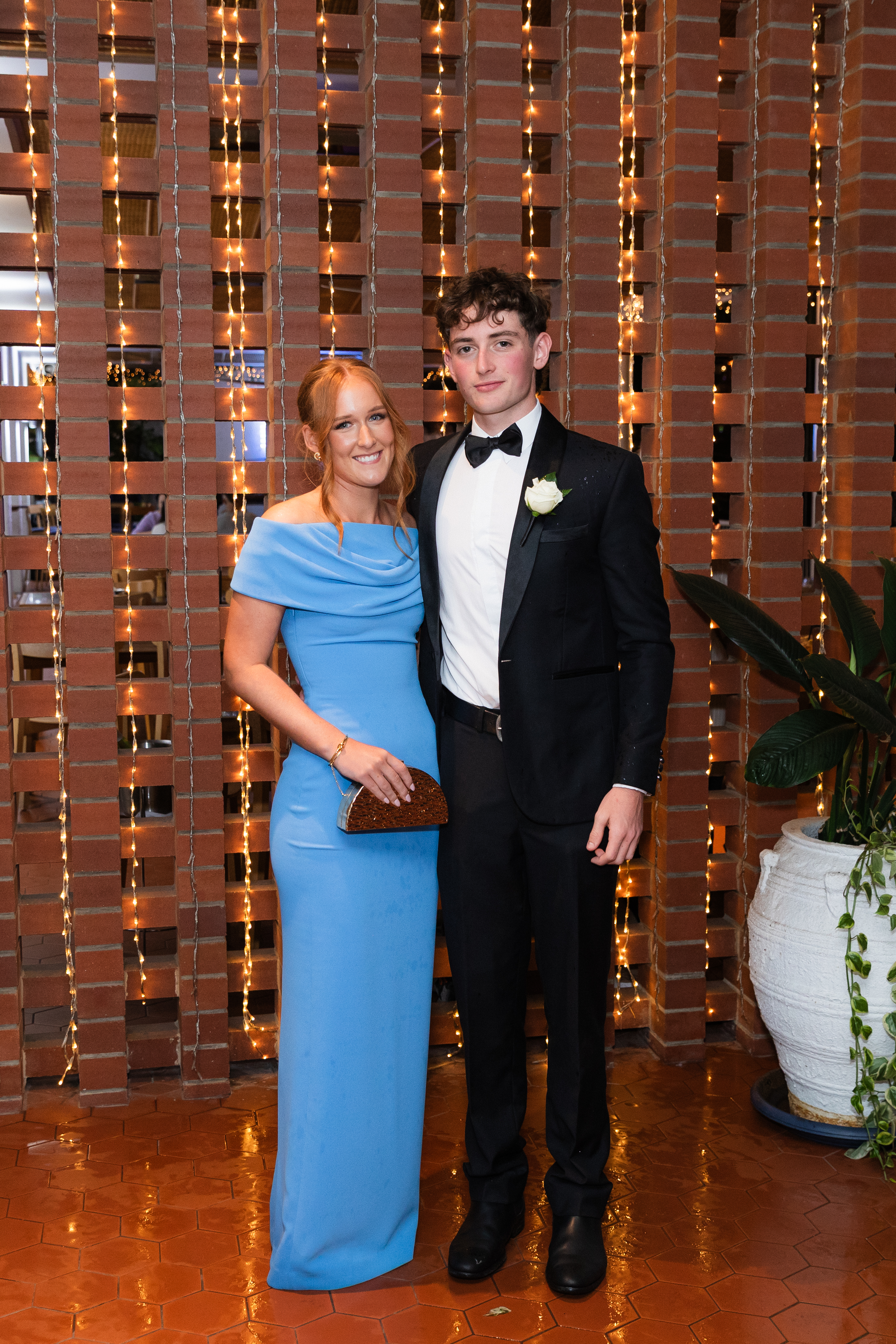 Two people standing side by side in formal evening wear in front of a brick wall with warm string lights, posing for a full‑length photo at an indoor event.