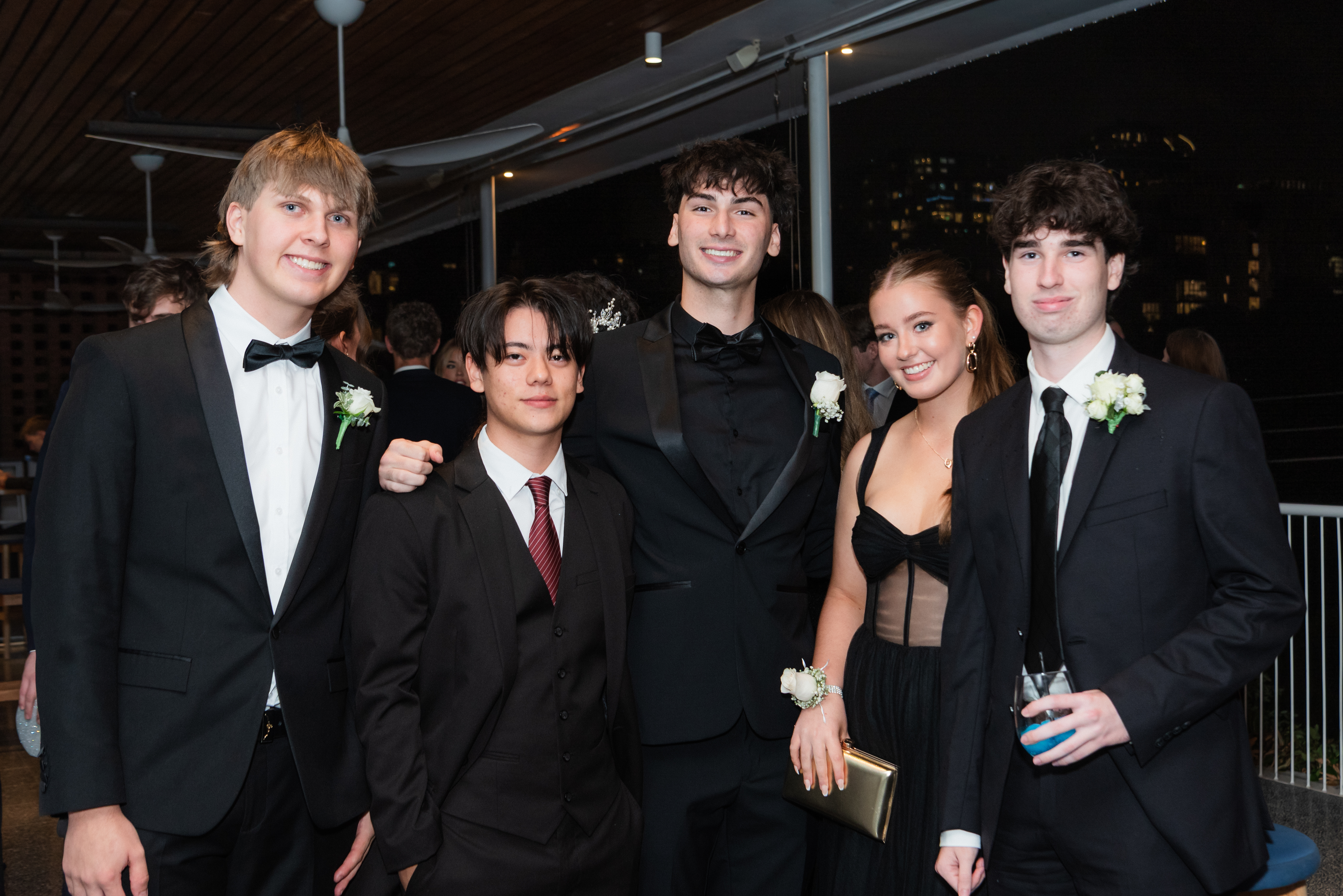 Five people dressed in formal evening wear standing together at an indoor event, with a crowd and city lights visible in the background.