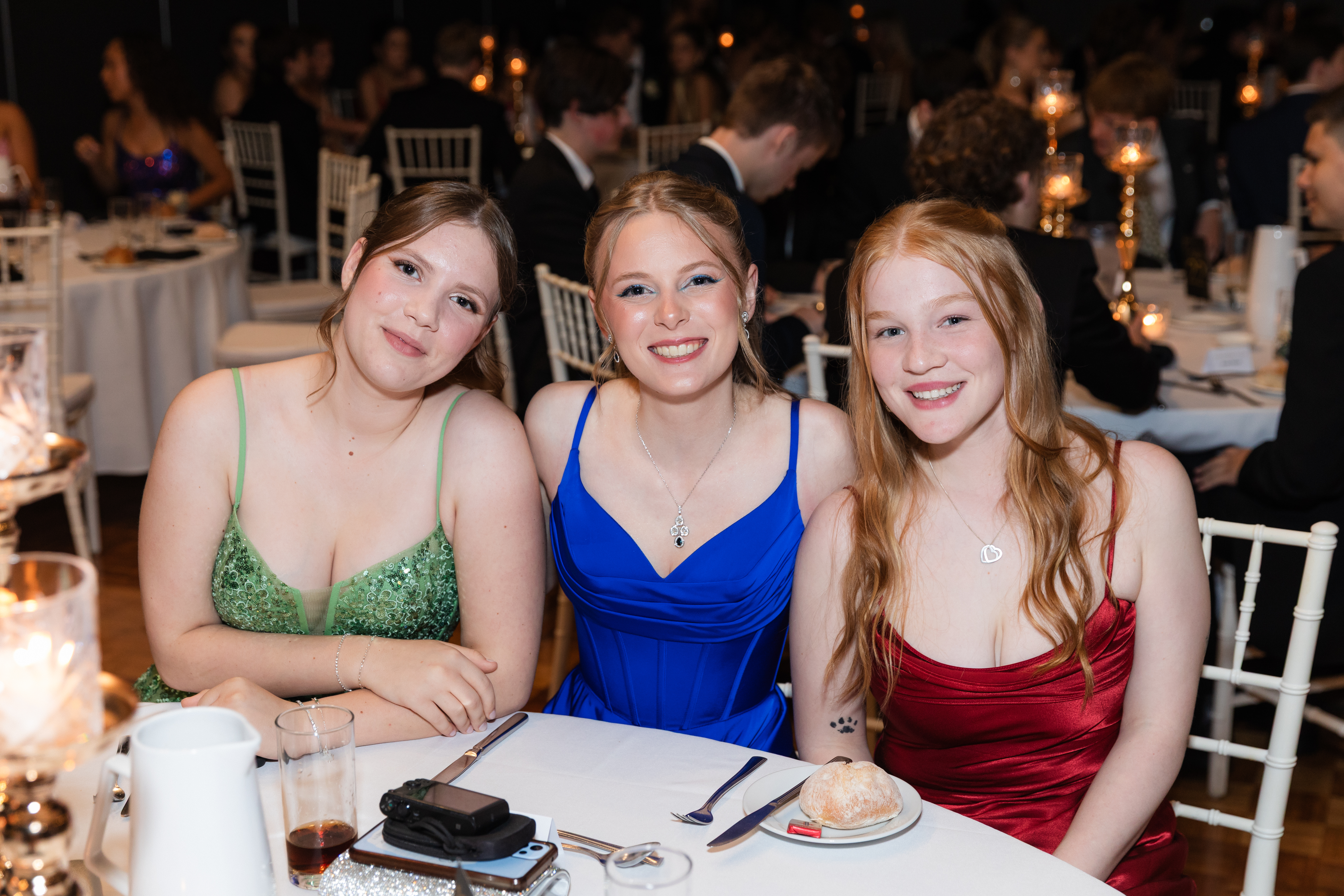 Three people seated at a round table in formal evening wear, posing for a photo at a candlelit indoor event with place settings and other tables in the background.
