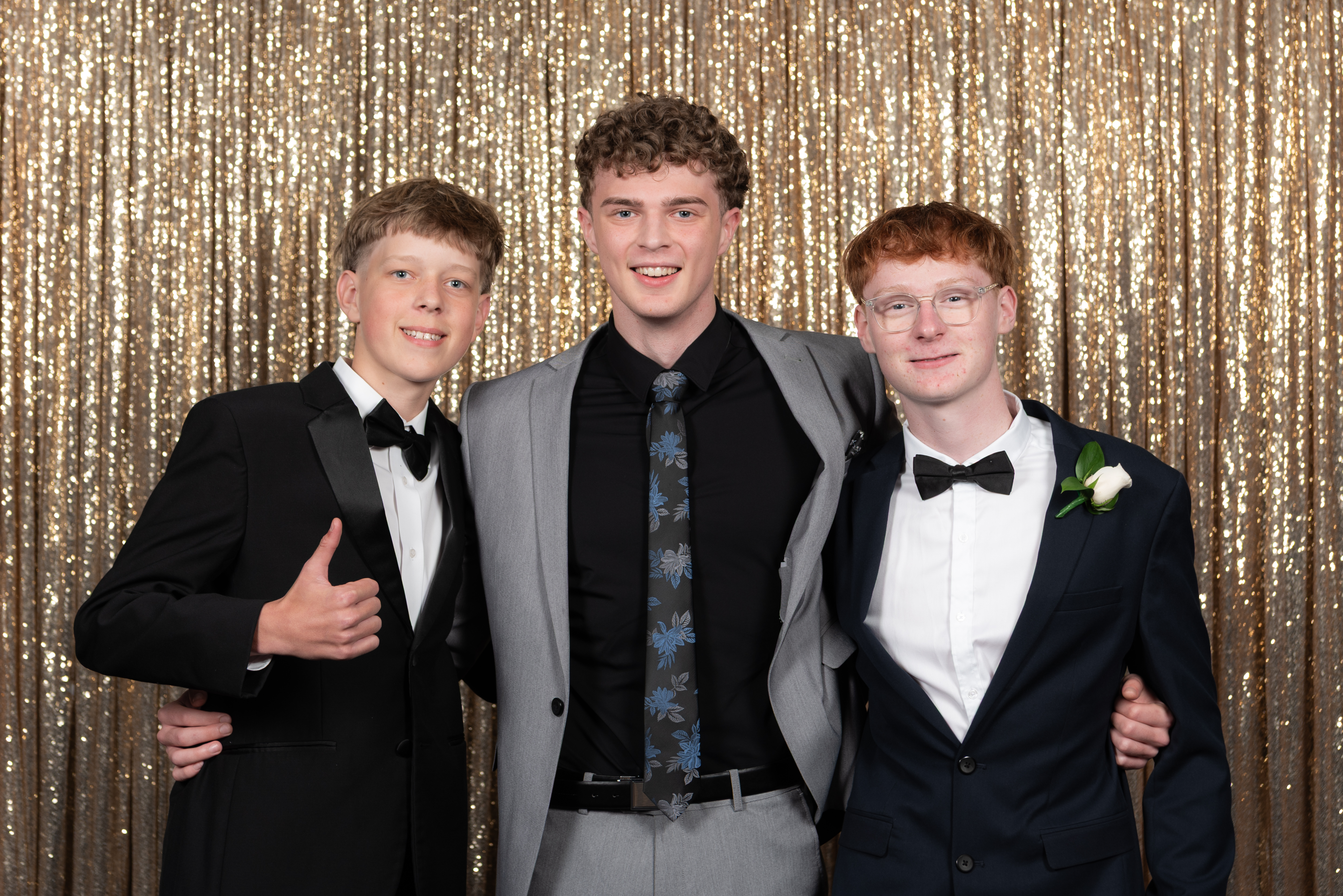 Three people standing close together in formal suits, posing in front of a gold sequin backdrop at an indoor evening event.