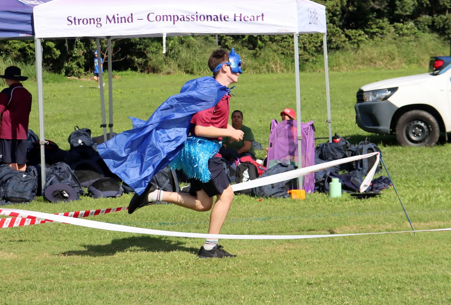 Student wearing a cape and mask runs across a marked grass field during a school carnival activity.