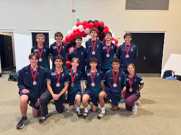 Group of students in sports uniforms wearing medals pose together indoors, with a red, black and white balloon arch behind them.