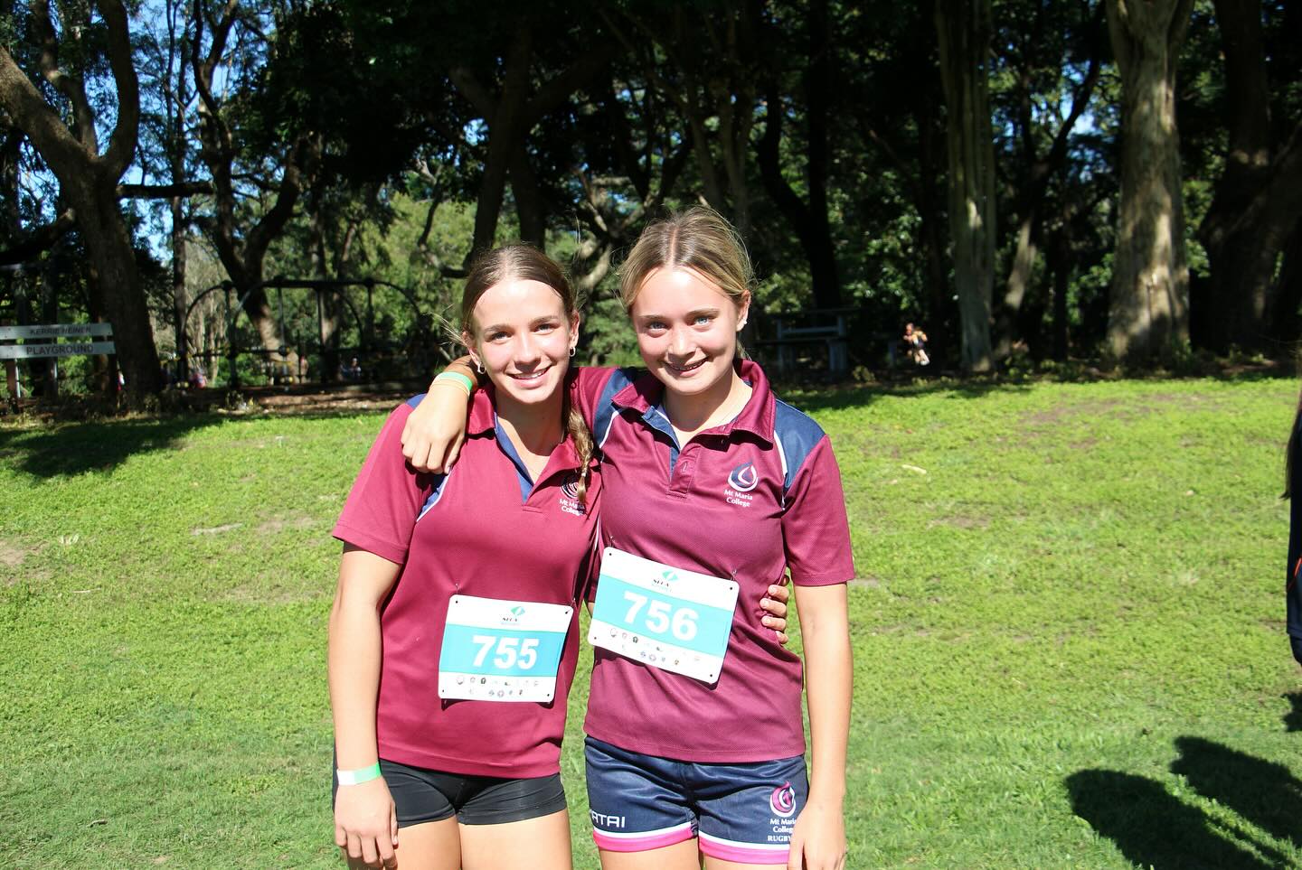 Two students in maroon school sports uniforms with race bibs stand together on a grassy field with arms around each other, with trees and spectators in the background.