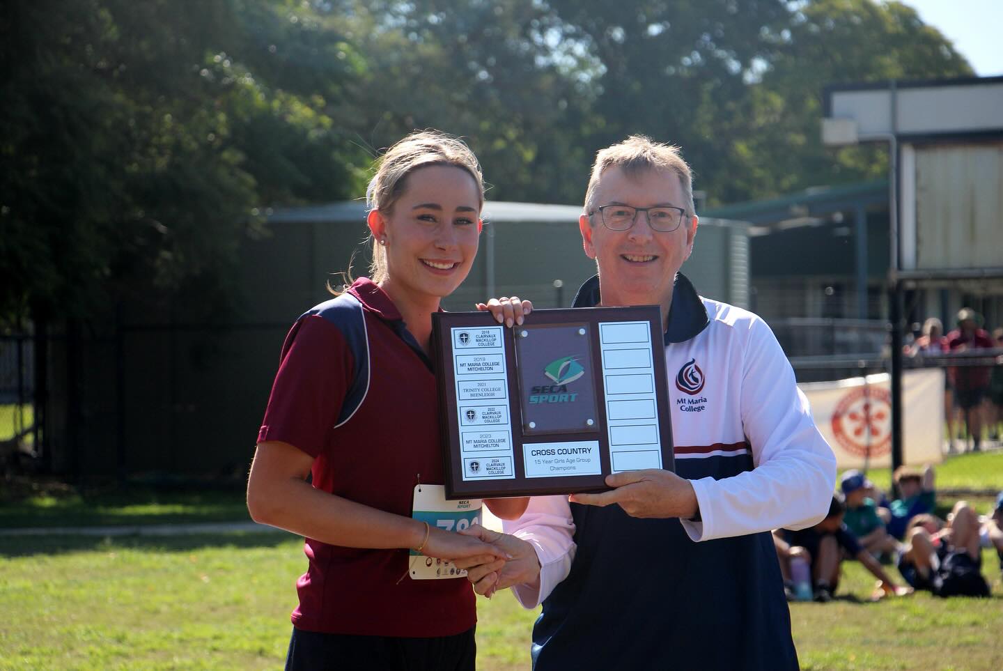 A student wearing a maroon sports uniform receives a cross‑country award plaque from a staff member on a grass field, with school buildings and seated spectators in the background.