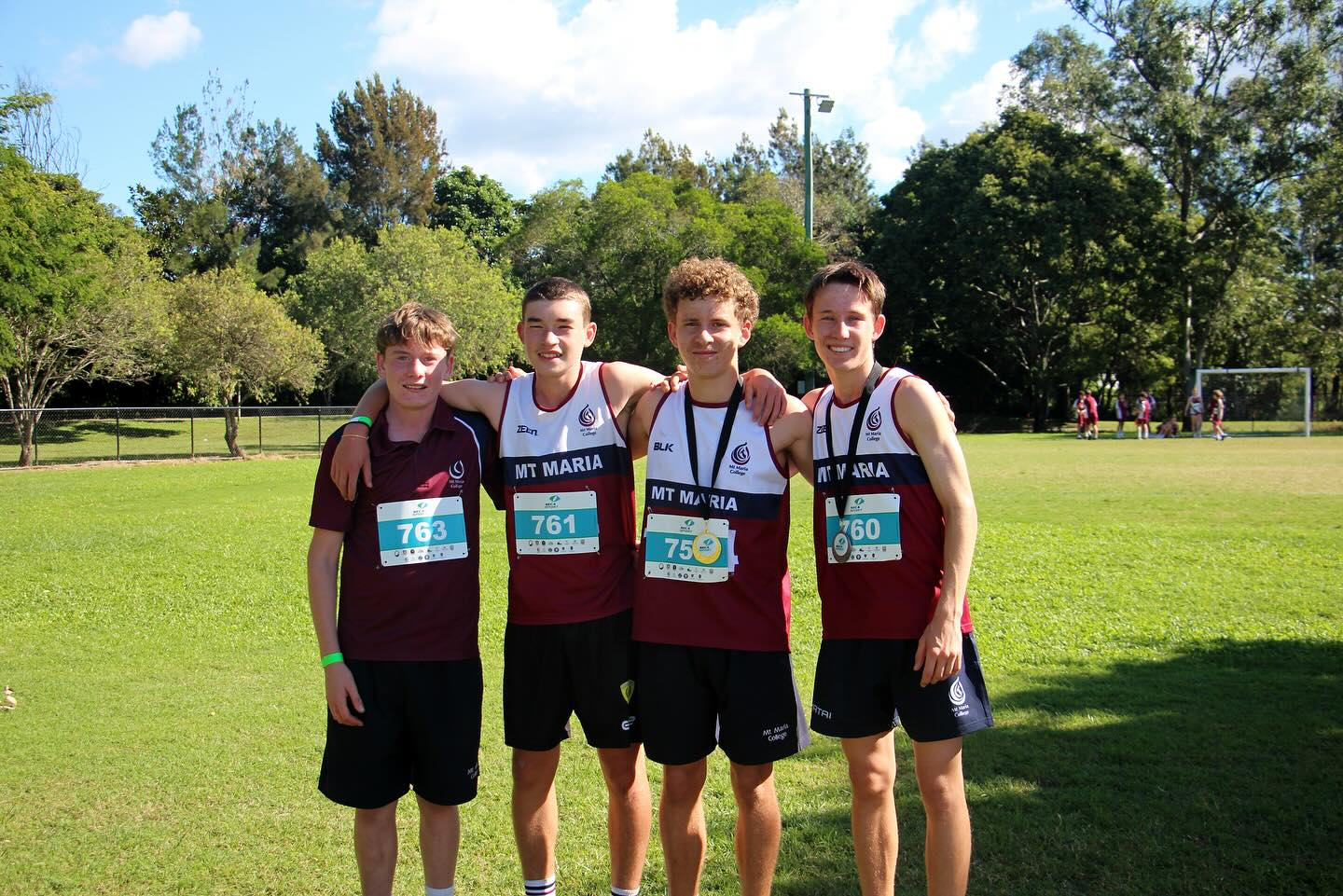 Four students wearing maroon school sports uniforms and race bibs stand side by side on a grassy field, with trees and other participants visible in the background.