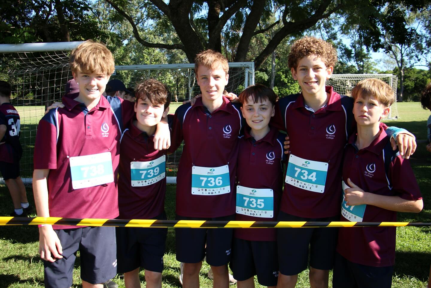 Six students wearing maroon school sports uniforms and race bibs stand together behind a course barrier on a grassy field, with trees and other competitors in the background.