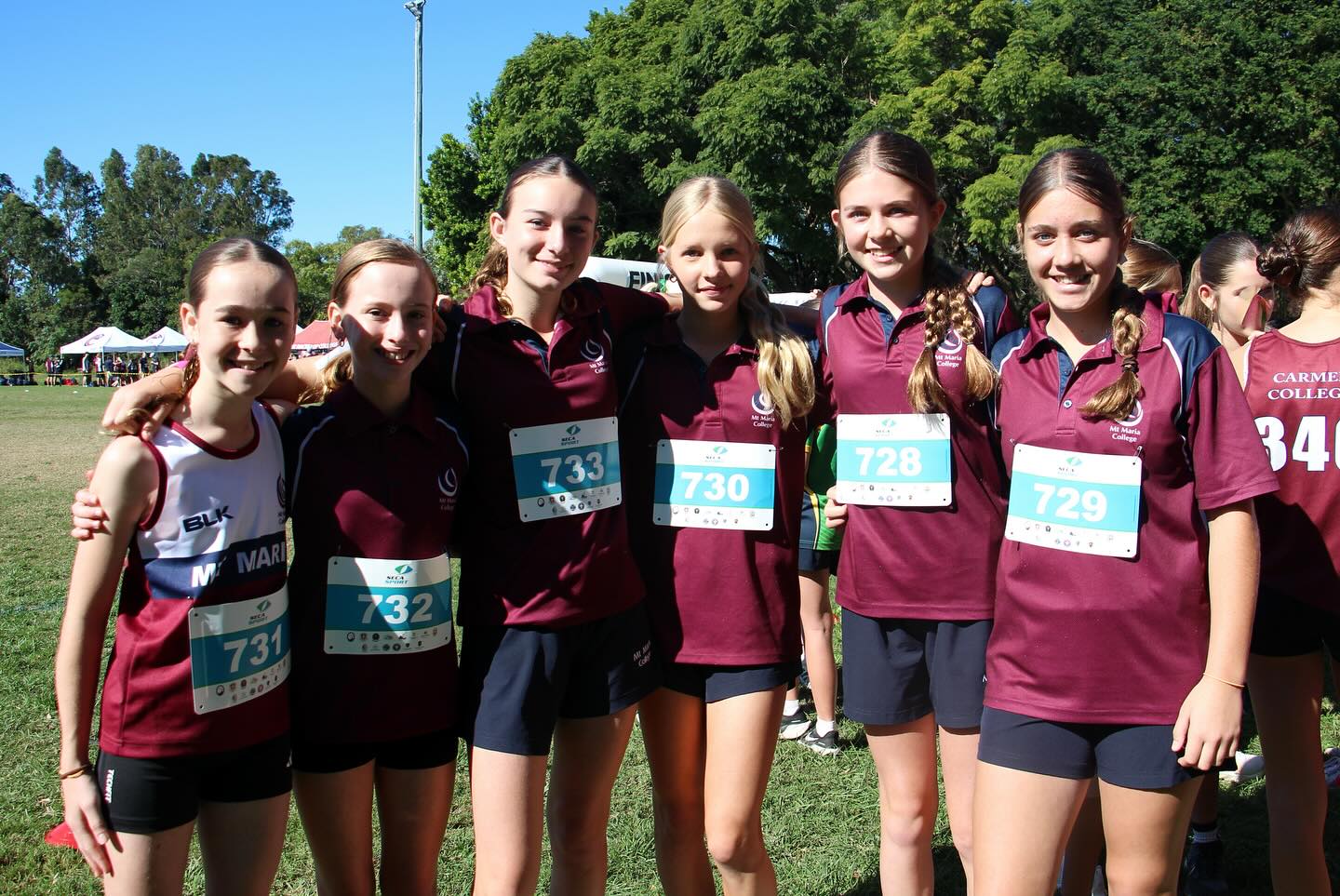 Six students wearing maroon school sports uniforms and numbered race bibs stand together on a grass field during a cross‑country event, with event tents and trees visible behind them.