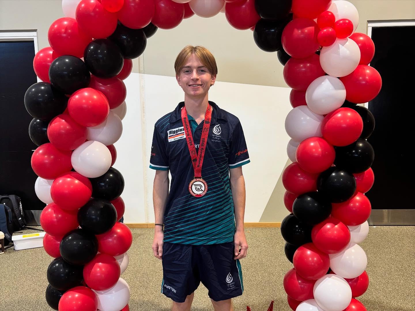 Student wearing a sports uniform and a medal stands beneath a red, black and white balloon arch inside a hall.