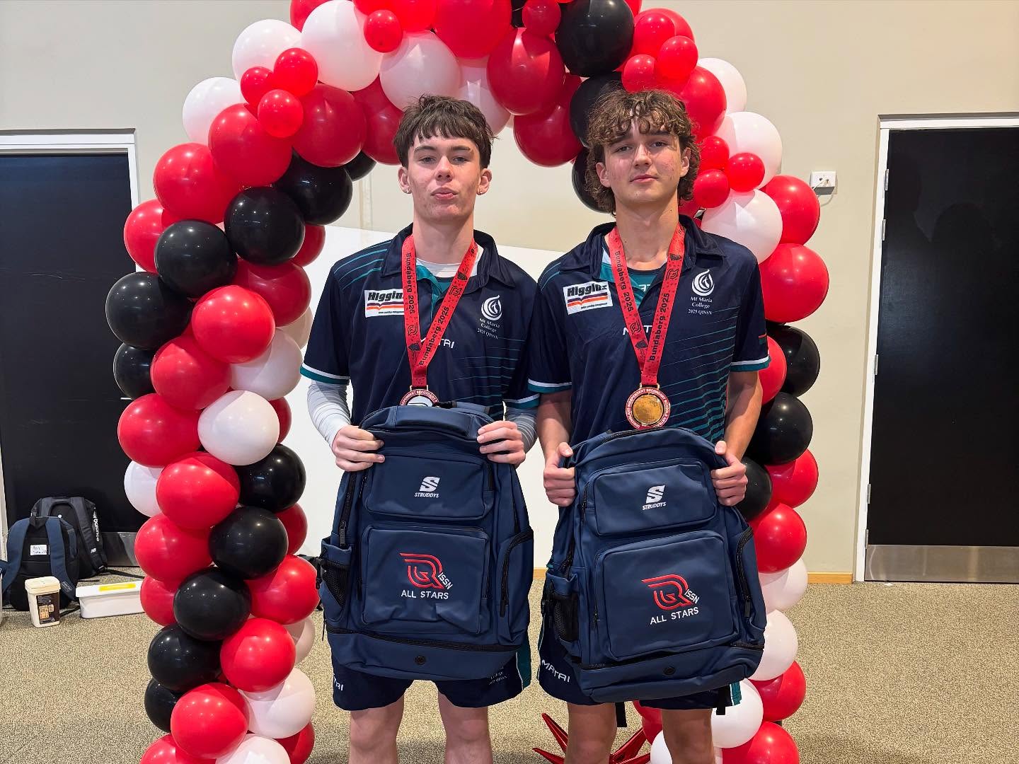 Two students in sports uniforms stand beneath a balloon arch holding sports backpacks, each wearing a medal.