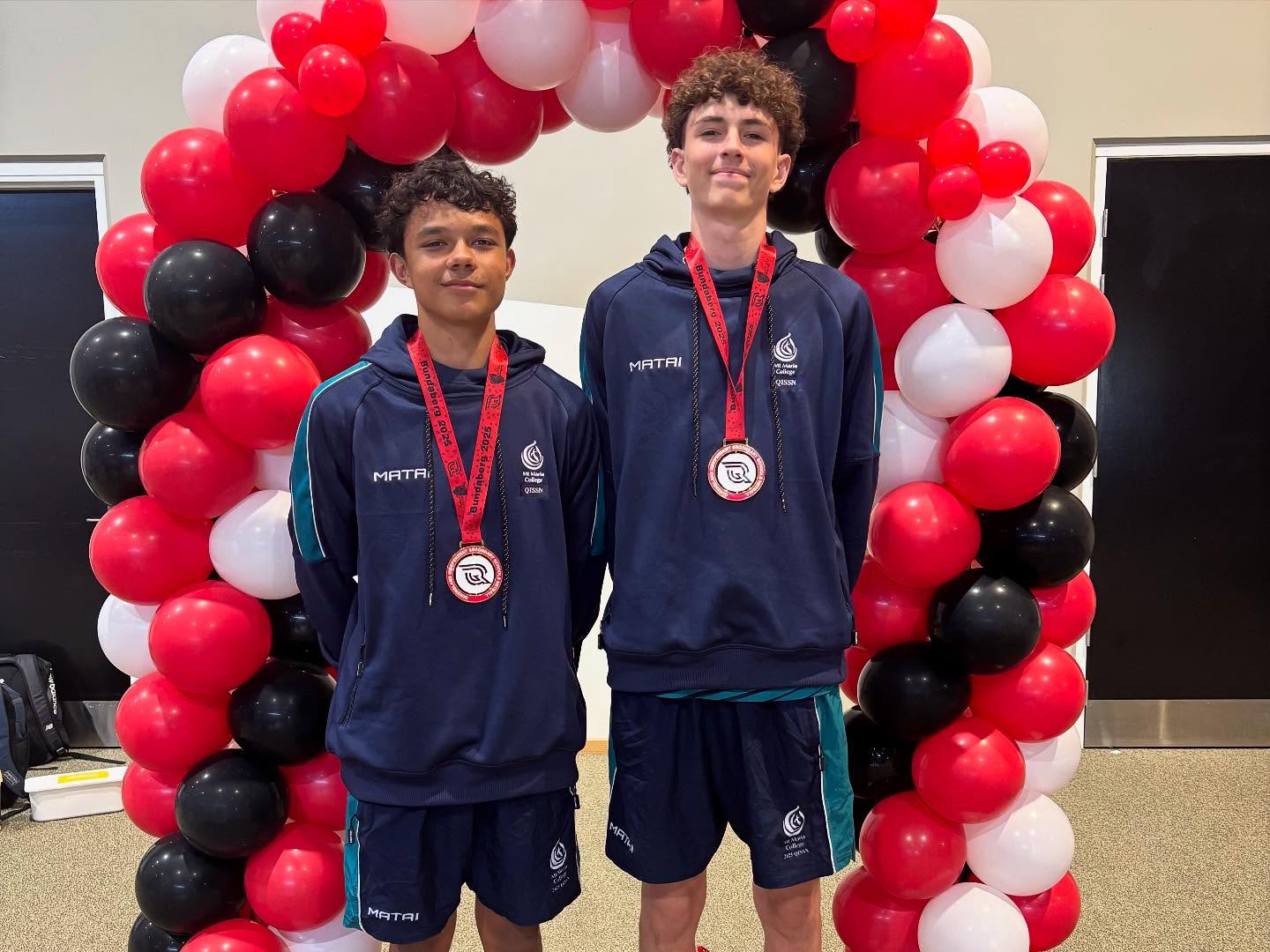 Two students in sports uniforms stand side by side beneath a red, black and white balloon arch, each wearing a medal.