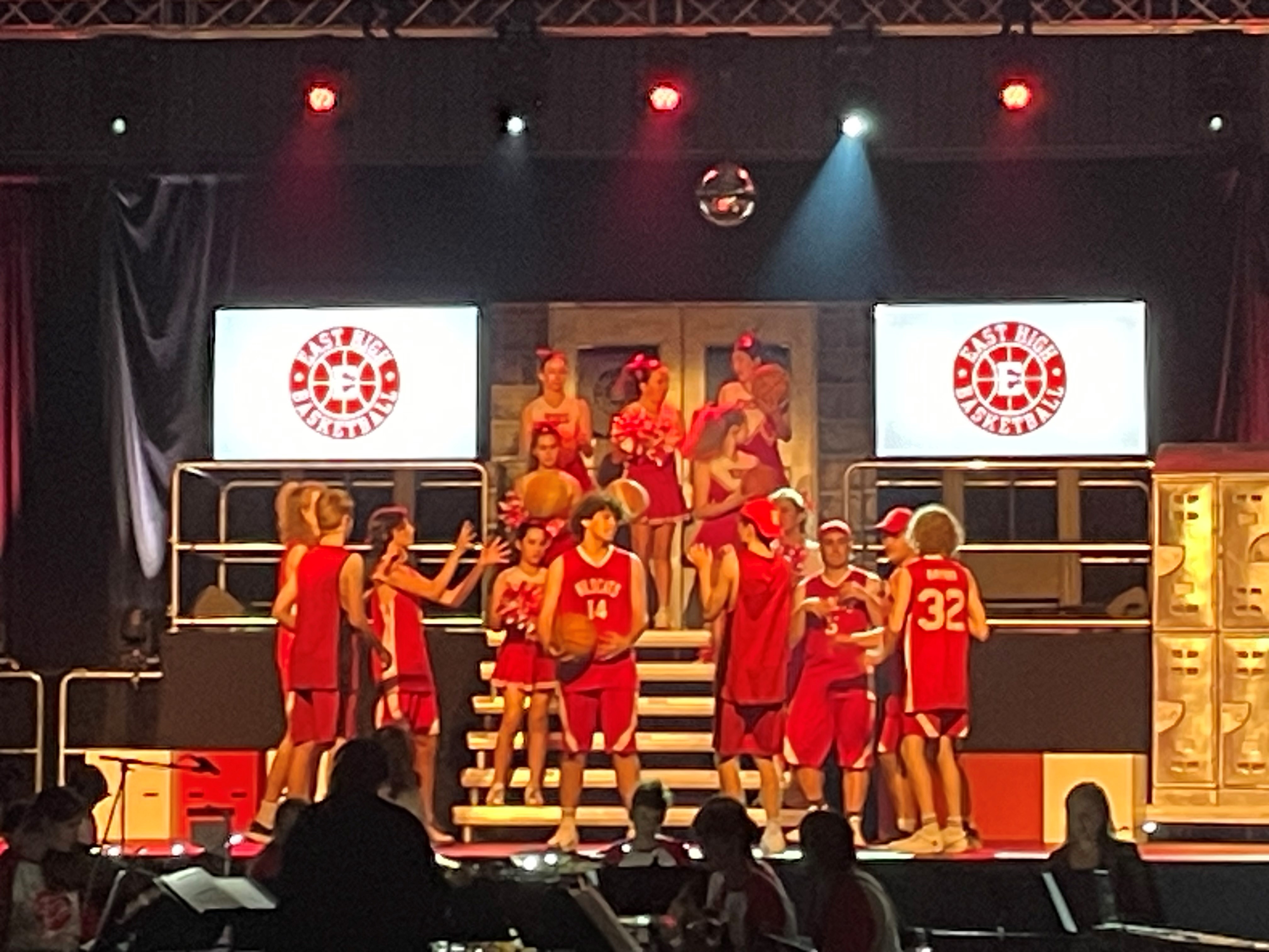 Students in matching red basketball uniforms standing and moving across a staged school performance set, with tiered steps, stage lighting and basketball-themed props.