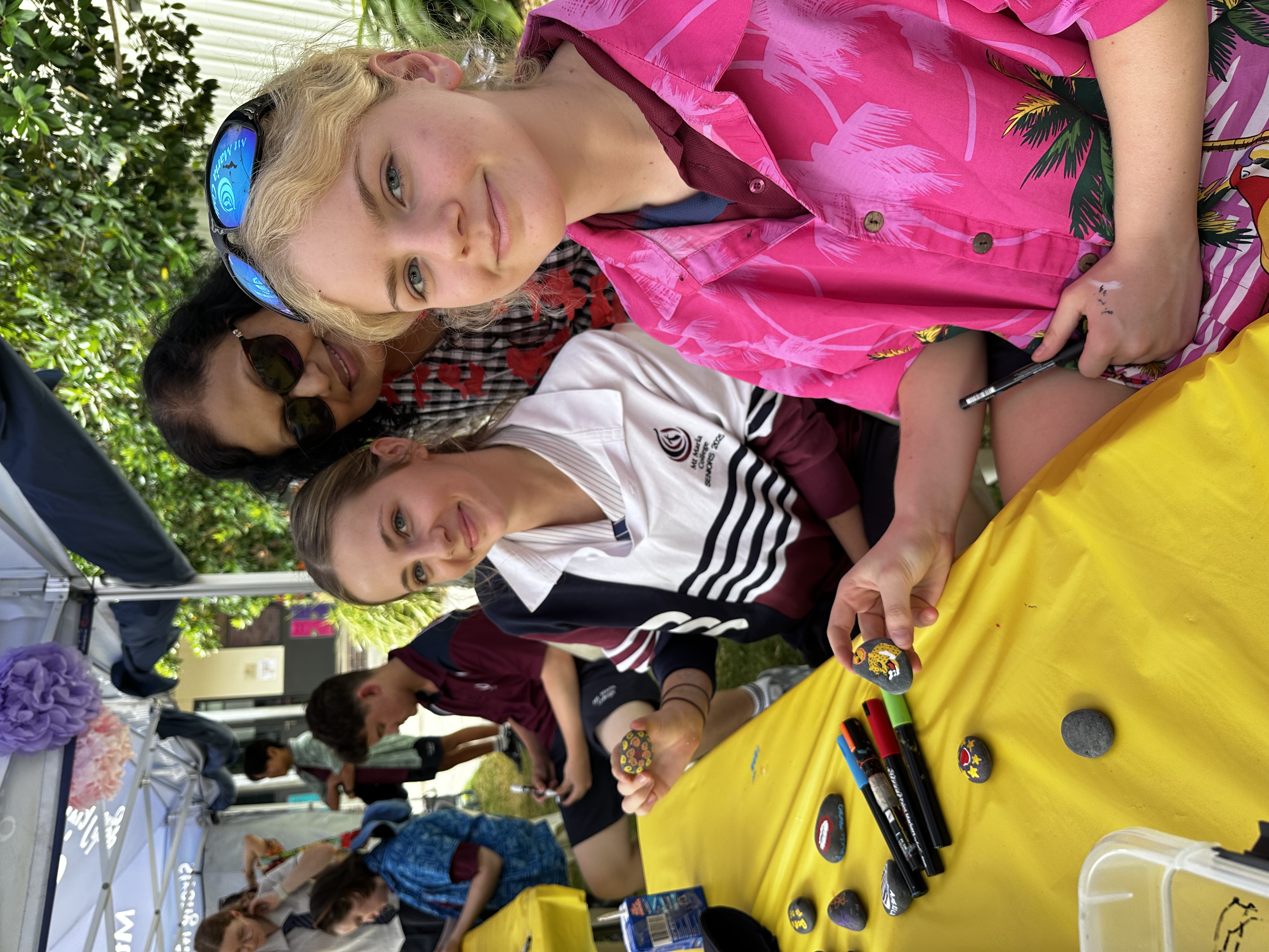 Two students sit at a yellow table decorating small black stones with markers as part of a school wellbeing activity. Painted stones, pens, and craft materials are spread across the table, with other students and activity stalls visible in the background.