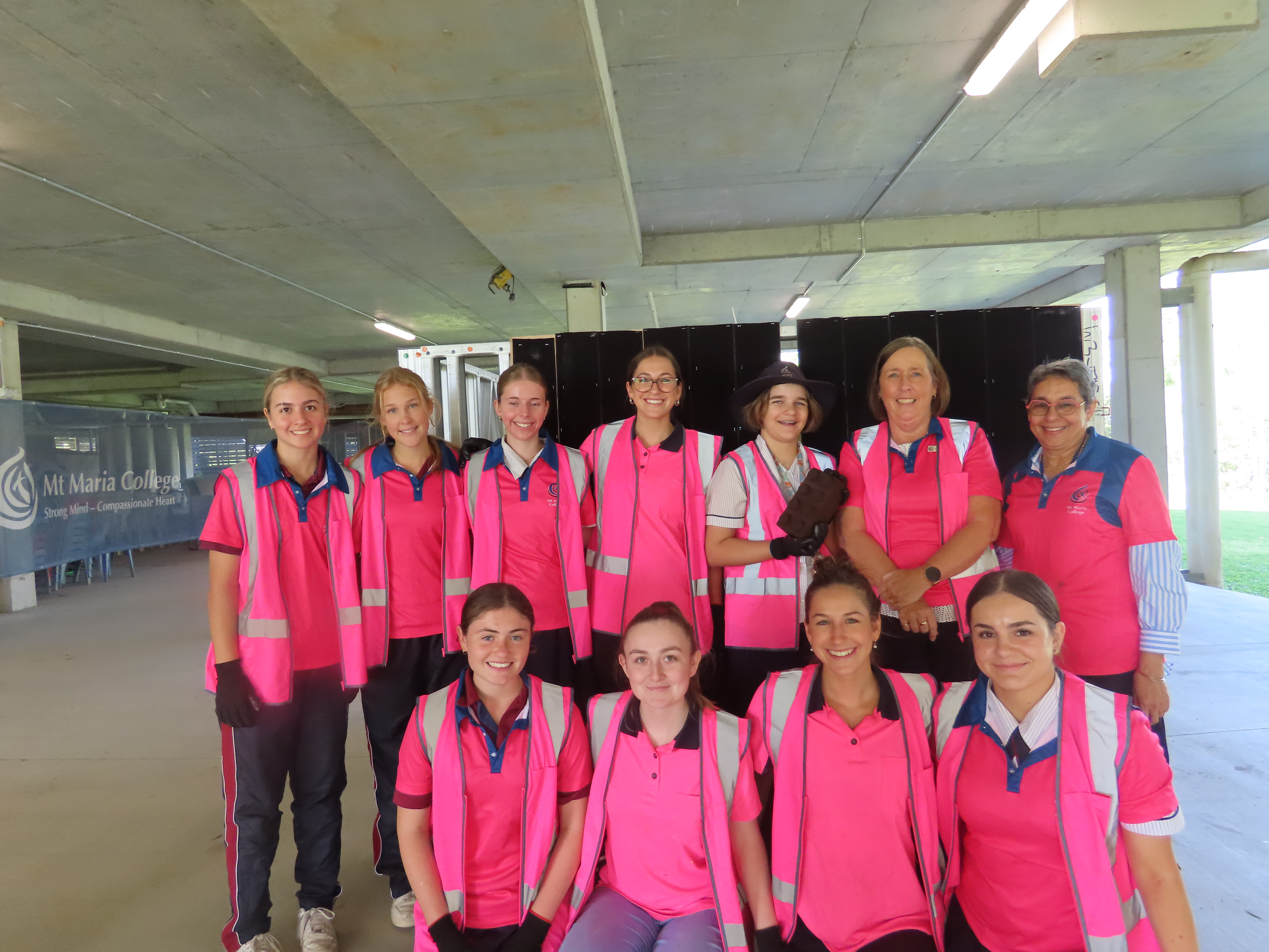 Students wearing high‑visibility pink work shirts stand with a secondary school student in uniform at a school construction site.