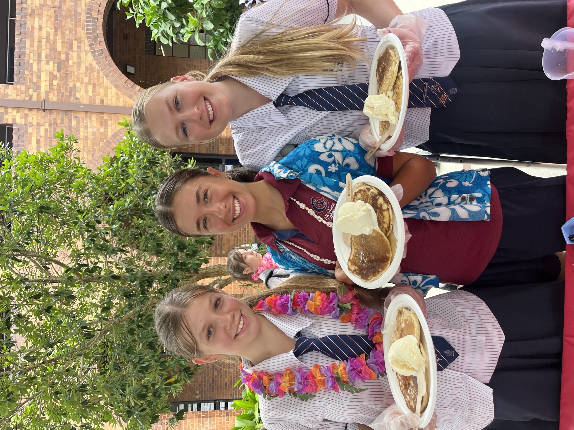 Three students in school uniform stand together holding plates of food during a school event on campus.