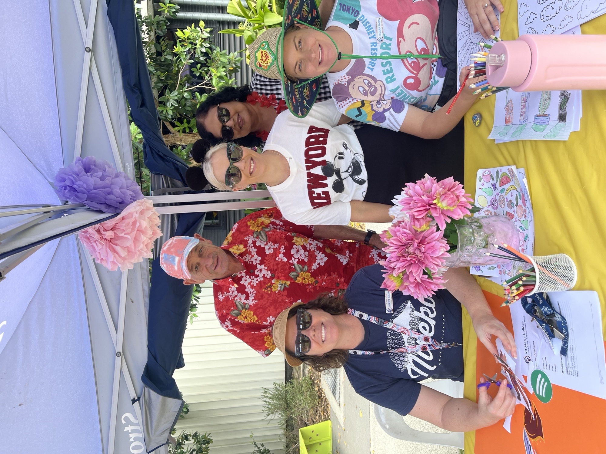 People sit and stand around a table under a marquee at a school wellbeing stall, colouring in artworks with pencils and felt‑tip pens. Bright paper flowers decorate the marquee, and colouring sheets, jars of pencils, and flowers are arranged on a yellow tablecloth.