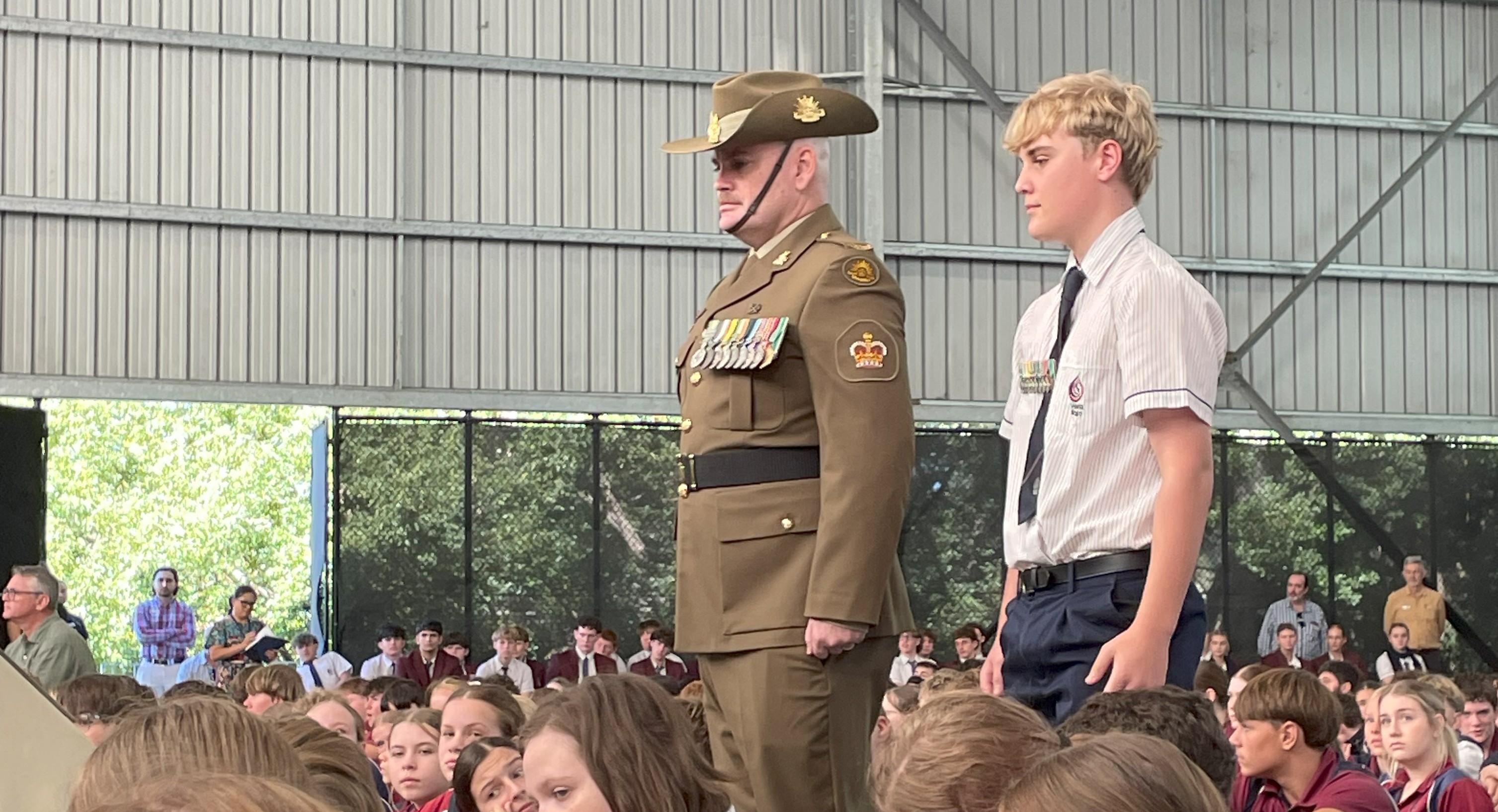 Uniformed defence personnel and a student stand at attention in front of a seated school assembly inside a covered hall.