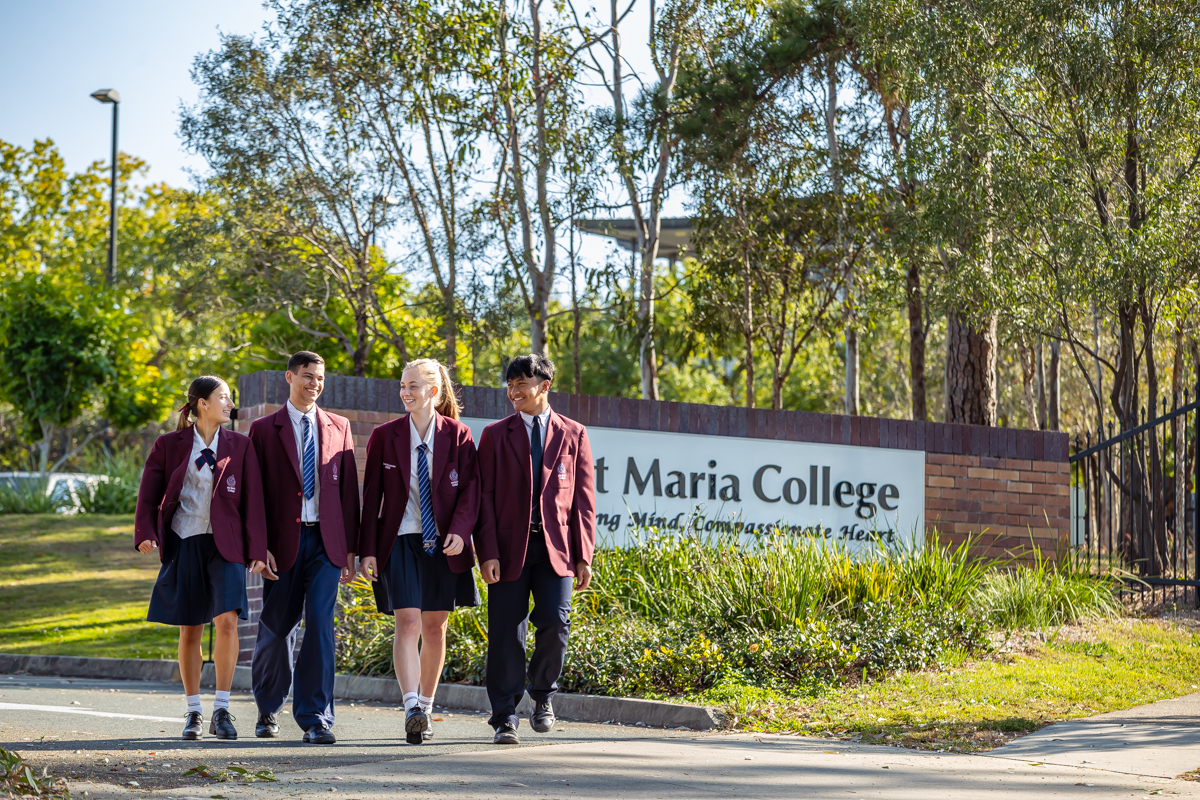 Four students in school uniforms walk together past the school entrance sign on a tree‑lined campus path.