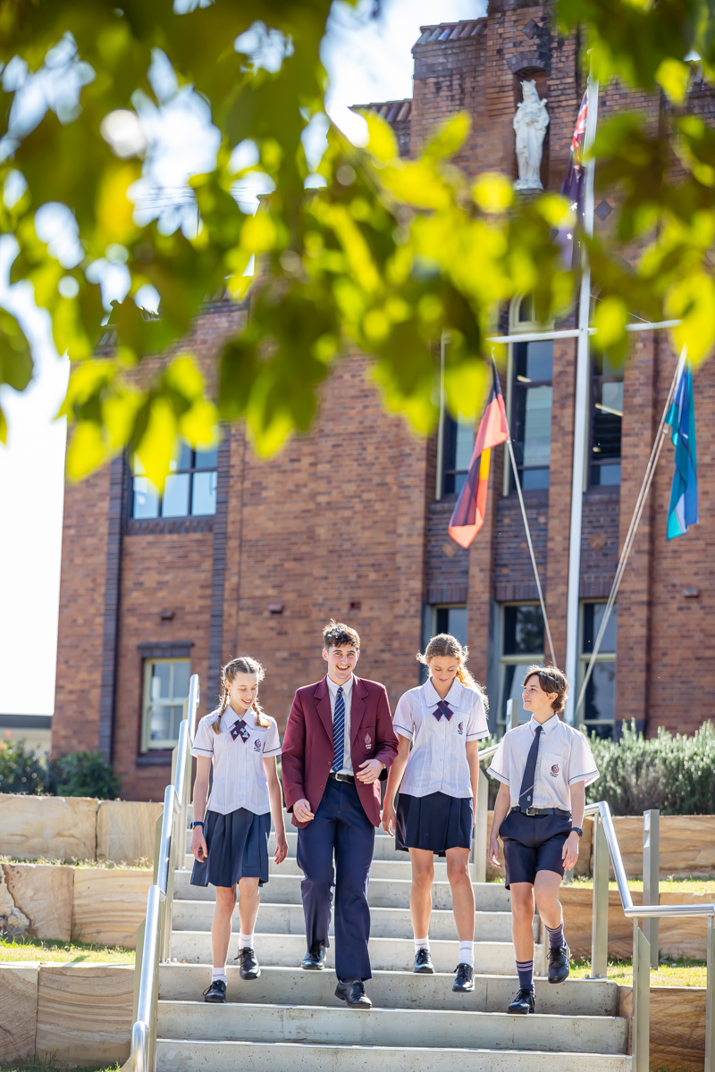 Students in school uniform walk down outdoor steps in front of a brick school building with flags displayed.