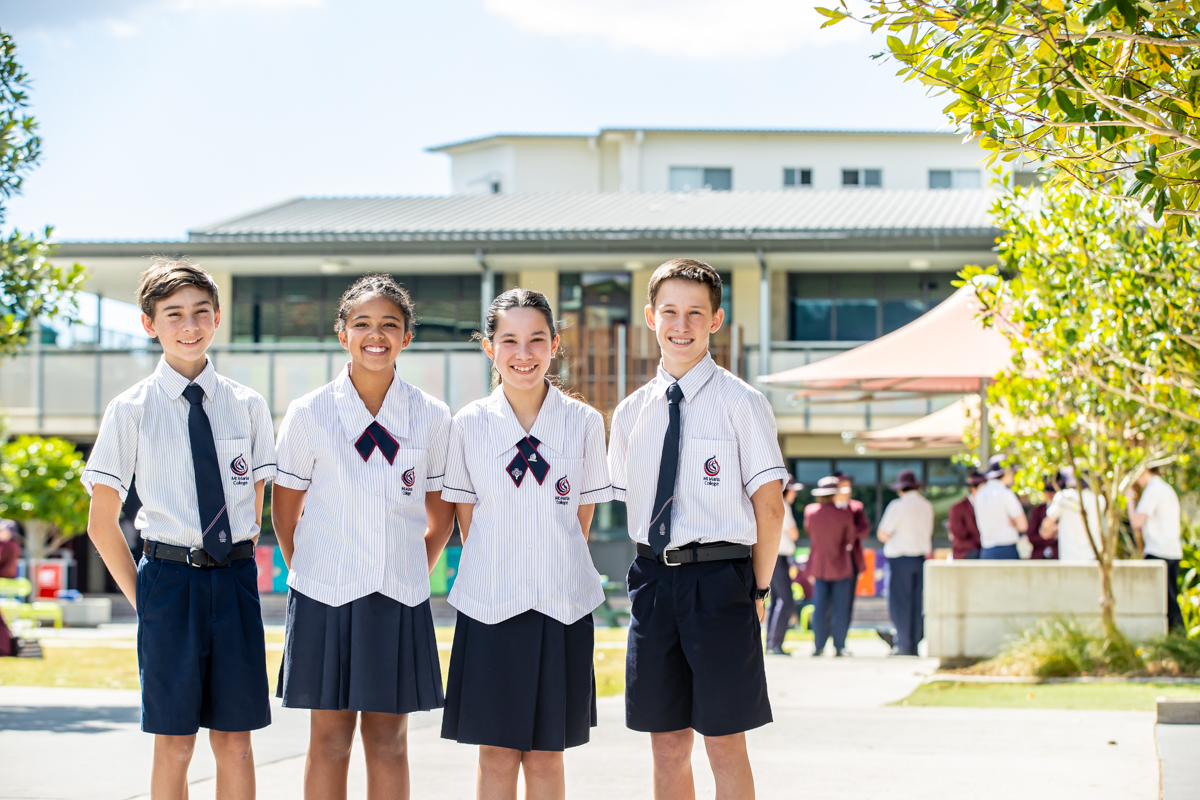 Four students stand side by side in school uniforms on a campus courtyard, with school buildings visible behind them.