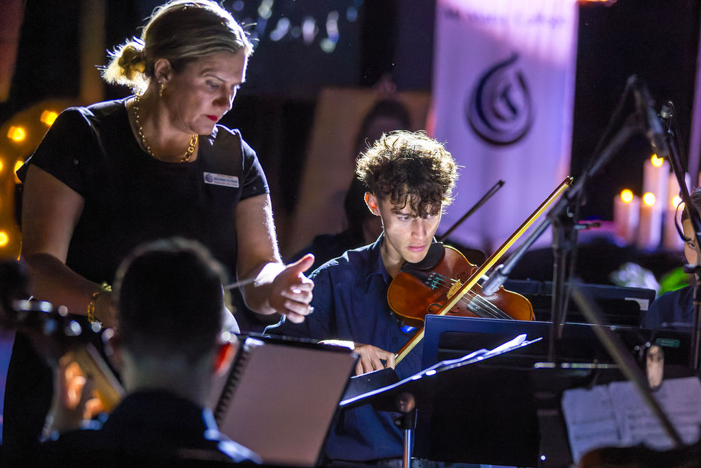 A teacher gestures toward sheet music while a student plays a violin during a supervised instrumental music performance or rehearsal.