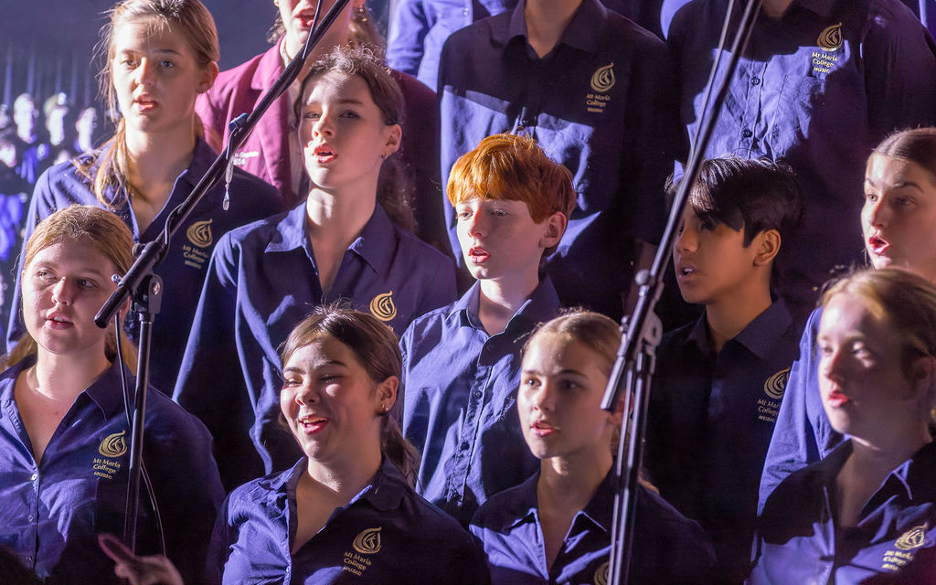 Students wearing school uniforms perform as part of a choir on stage, standing in rows and singing into microphones during a school performance.