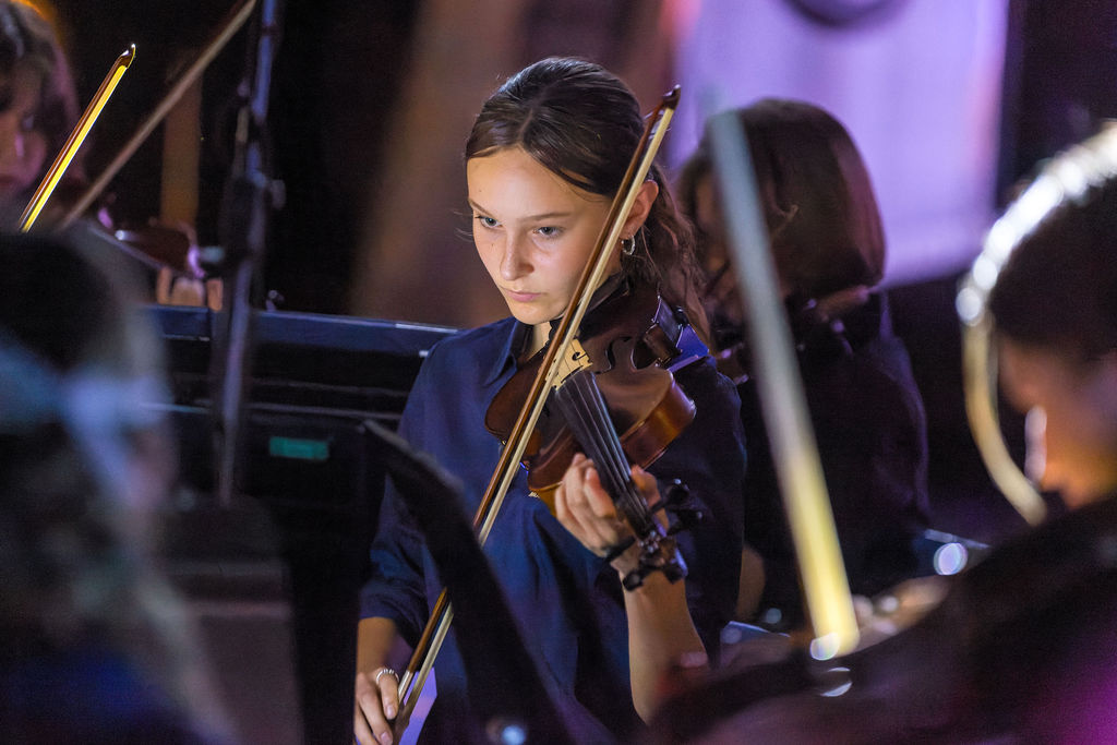 A student plays the violin while reading from sheet music during a school orchestral performance or rehearsal.