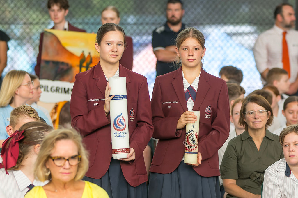 Two students in school uniform stand holding large ceremonial candles during a school gathering, with other students and adults seated behind them.