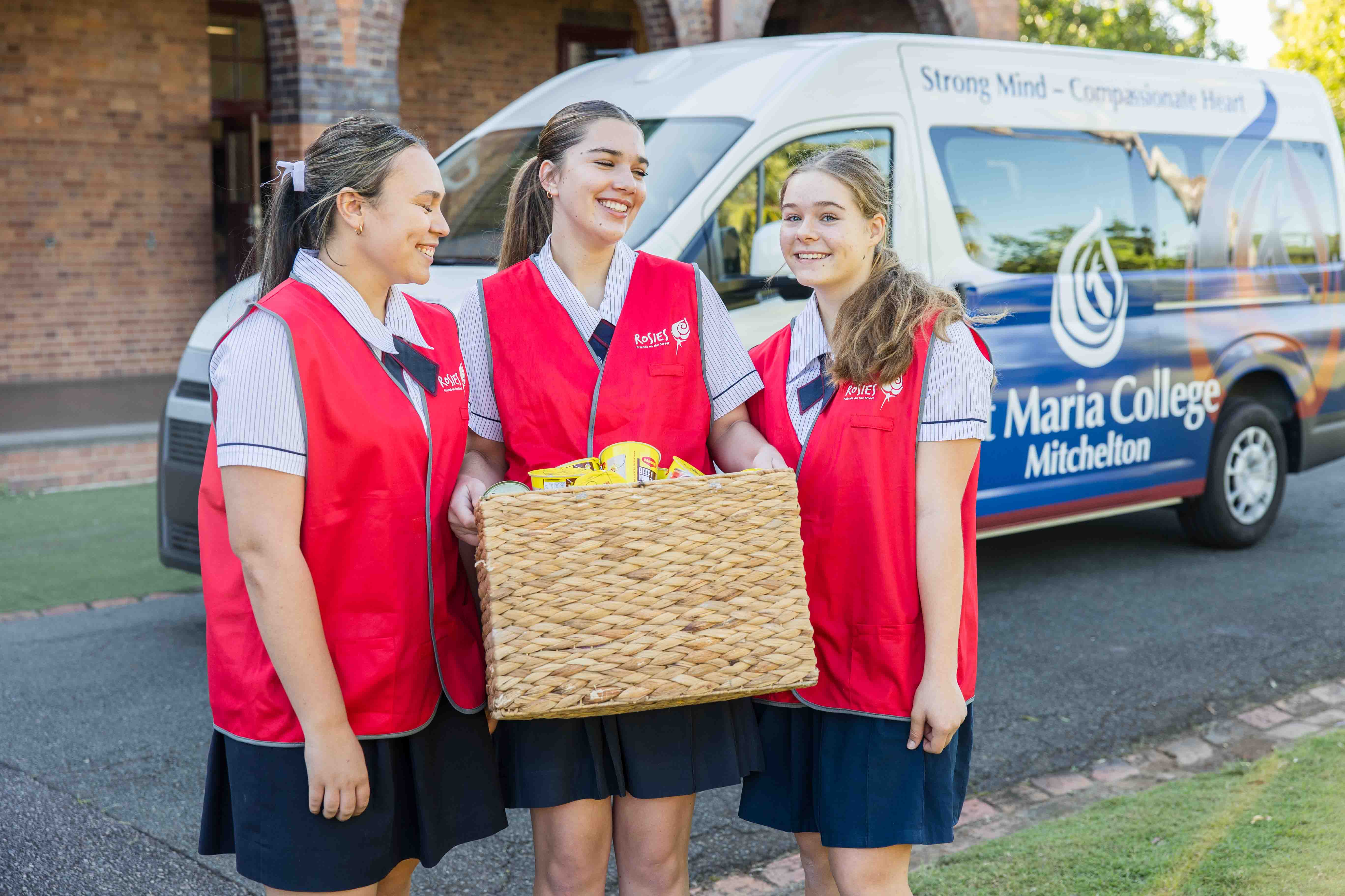 Three student volunteers wearing red vests hold a wicker basket in front of a school van on campus.