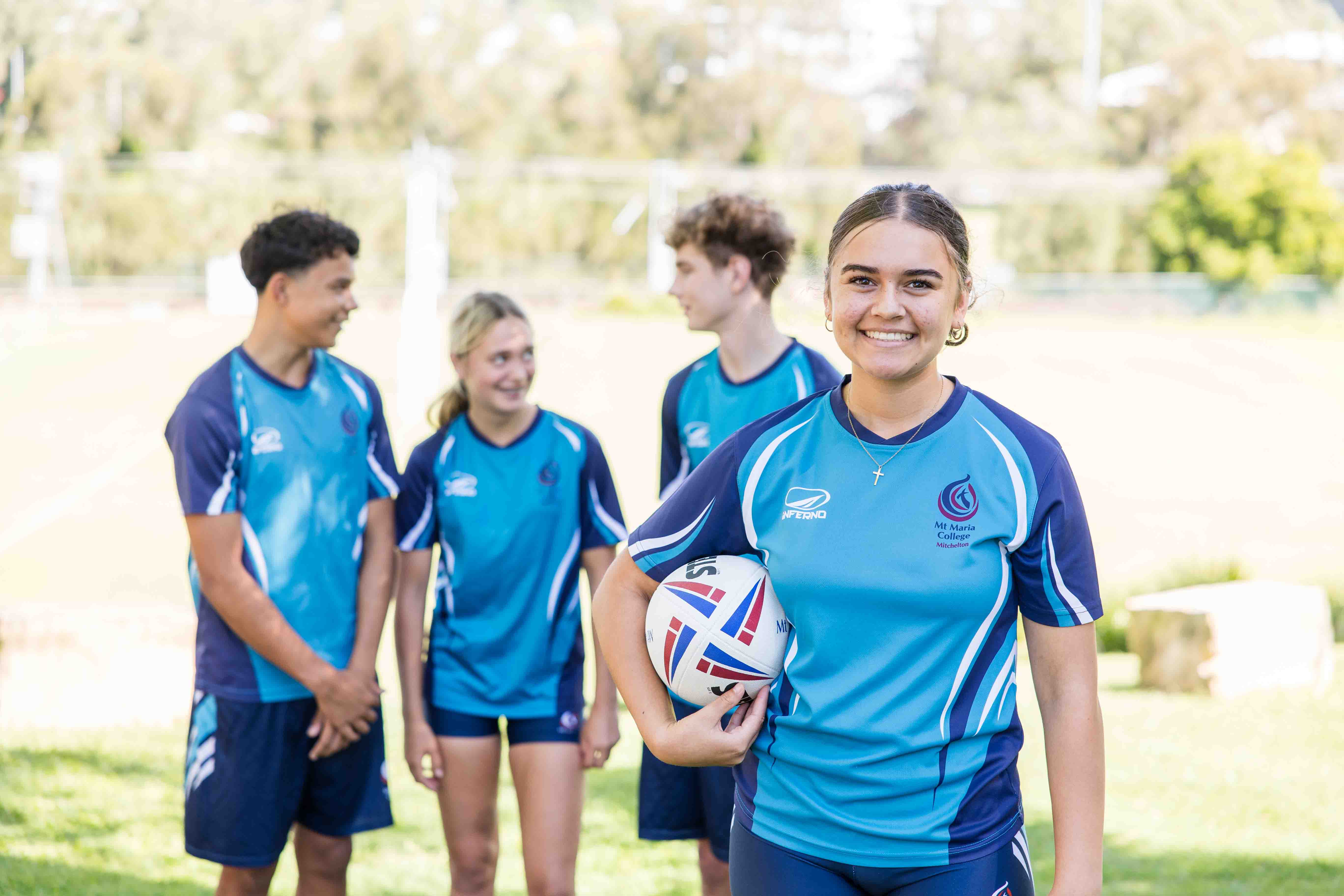 Student in a school sports uniform holding a rugby ball in the foreground, with three other students in matching uniforms standing behind on a grassy sports field.
