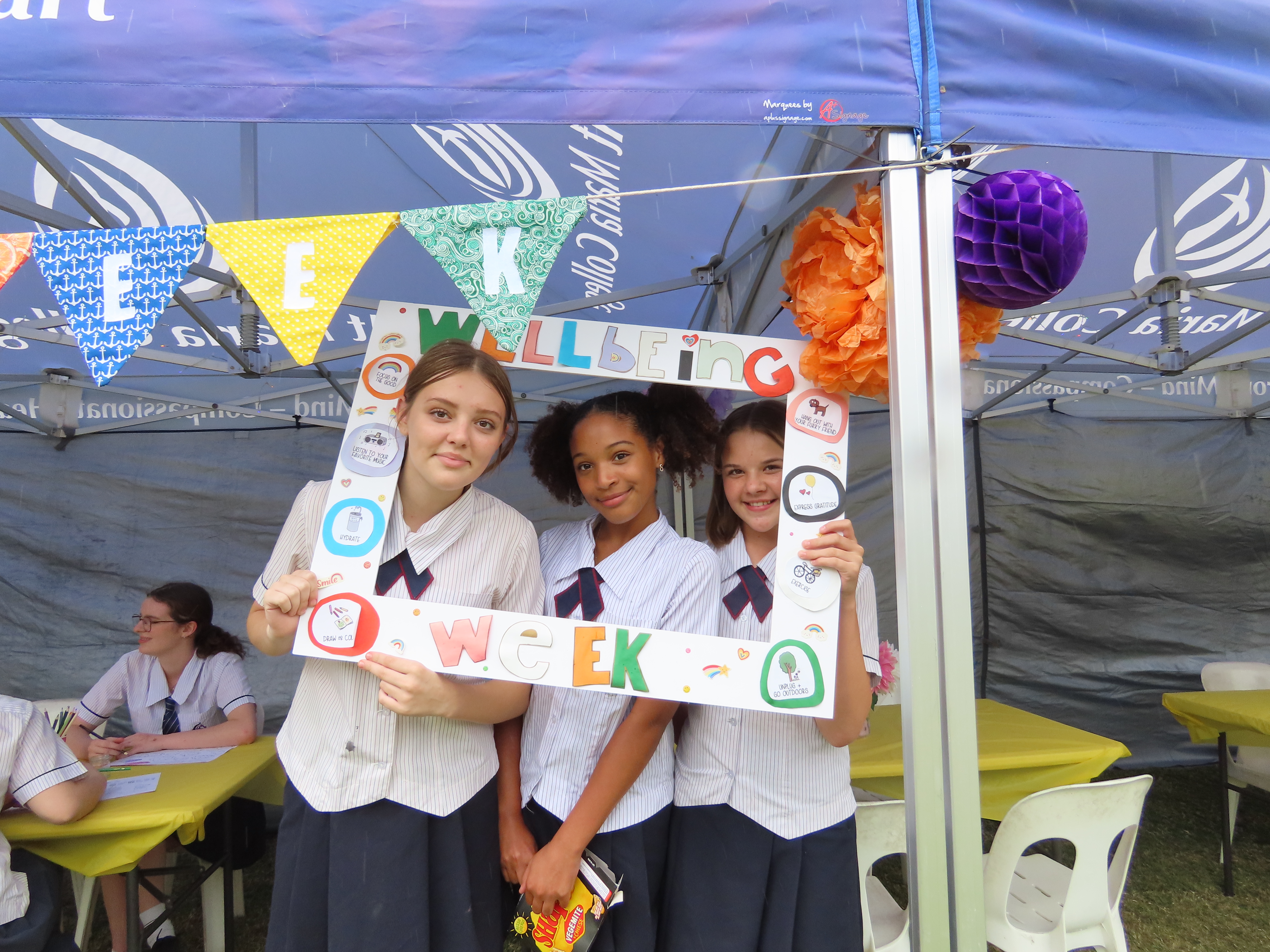 Three students in school uniform stand under a marquee holding a large decorative photo frame labelled “Wellbeing Week”. The frame includes colourful icons related to wellbeing and is displayed in front of a school wellbeing activity stall with bunting and tables behind them.
